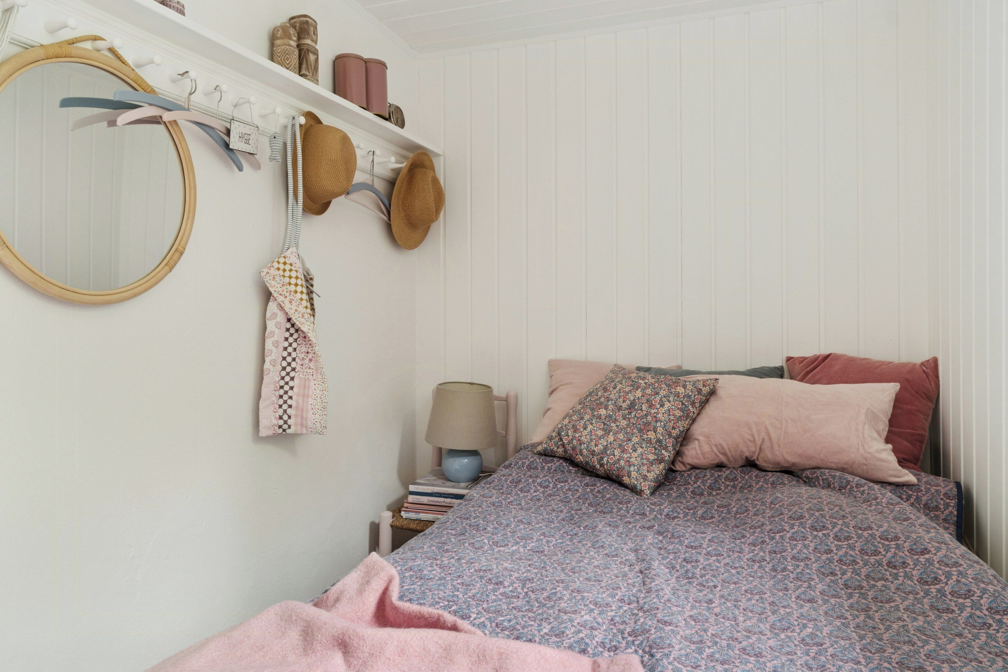 A bedroom with a bed, pillows, a hanging lamp, a mirror, and a shelf with hats and books.