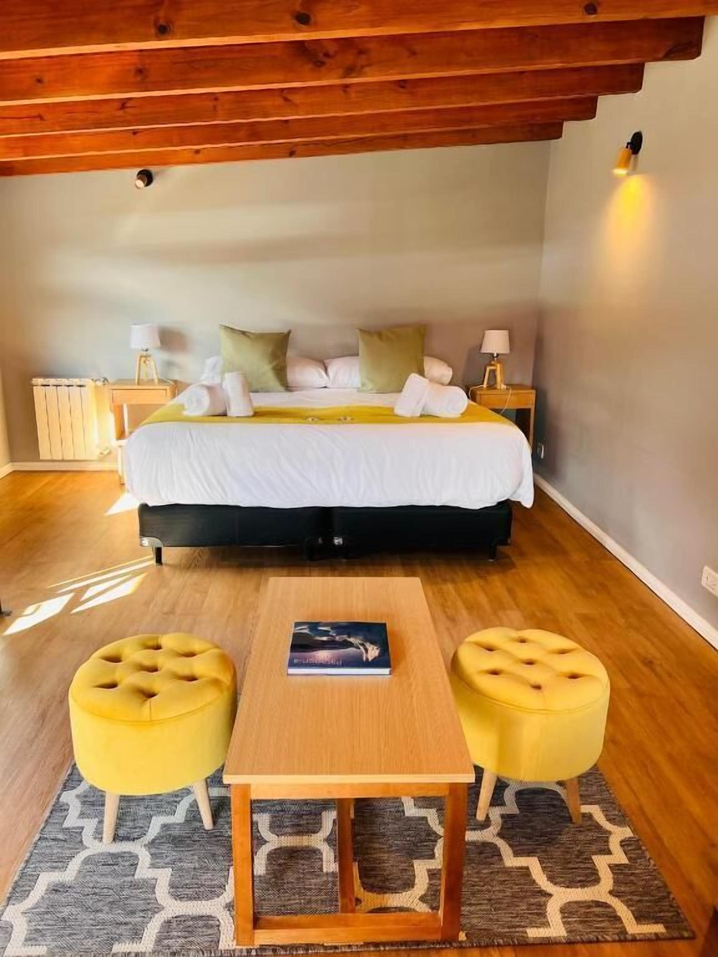 Un dormitorio con una cama, lámparas de noche, una mesa y dos otomanas amarillas.