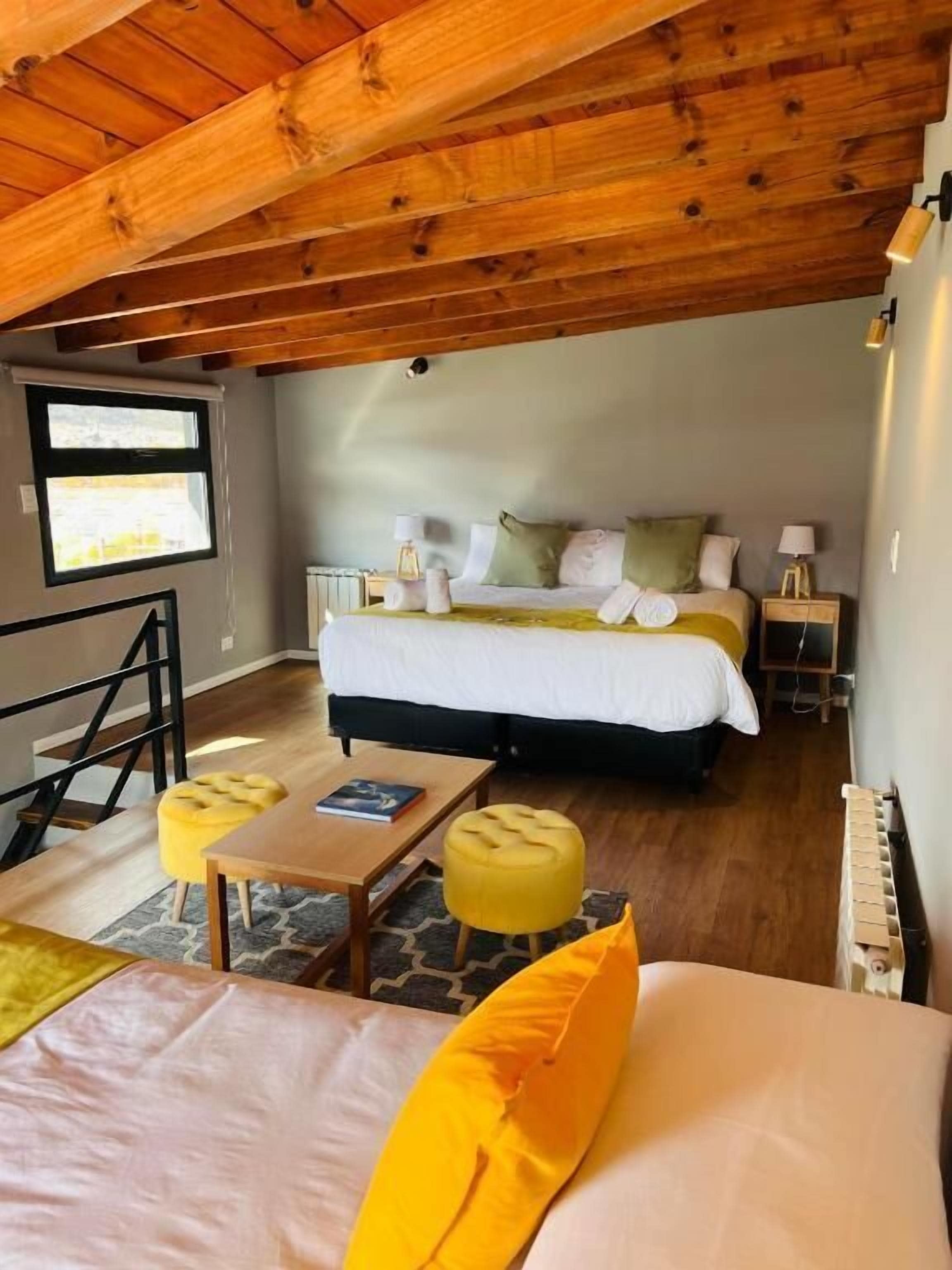 Quarto com uma cama, duas mesas de cabeceira, uma janela e um lance de escadas, além de um banquinho amarelo.