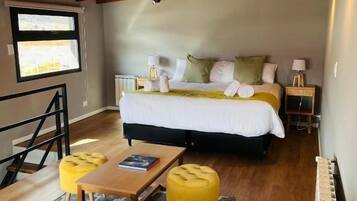 Quarto com uma cama, duas mesas de cabeceira, uma janela e um lance de escadas, além de um banquinho amarelo.