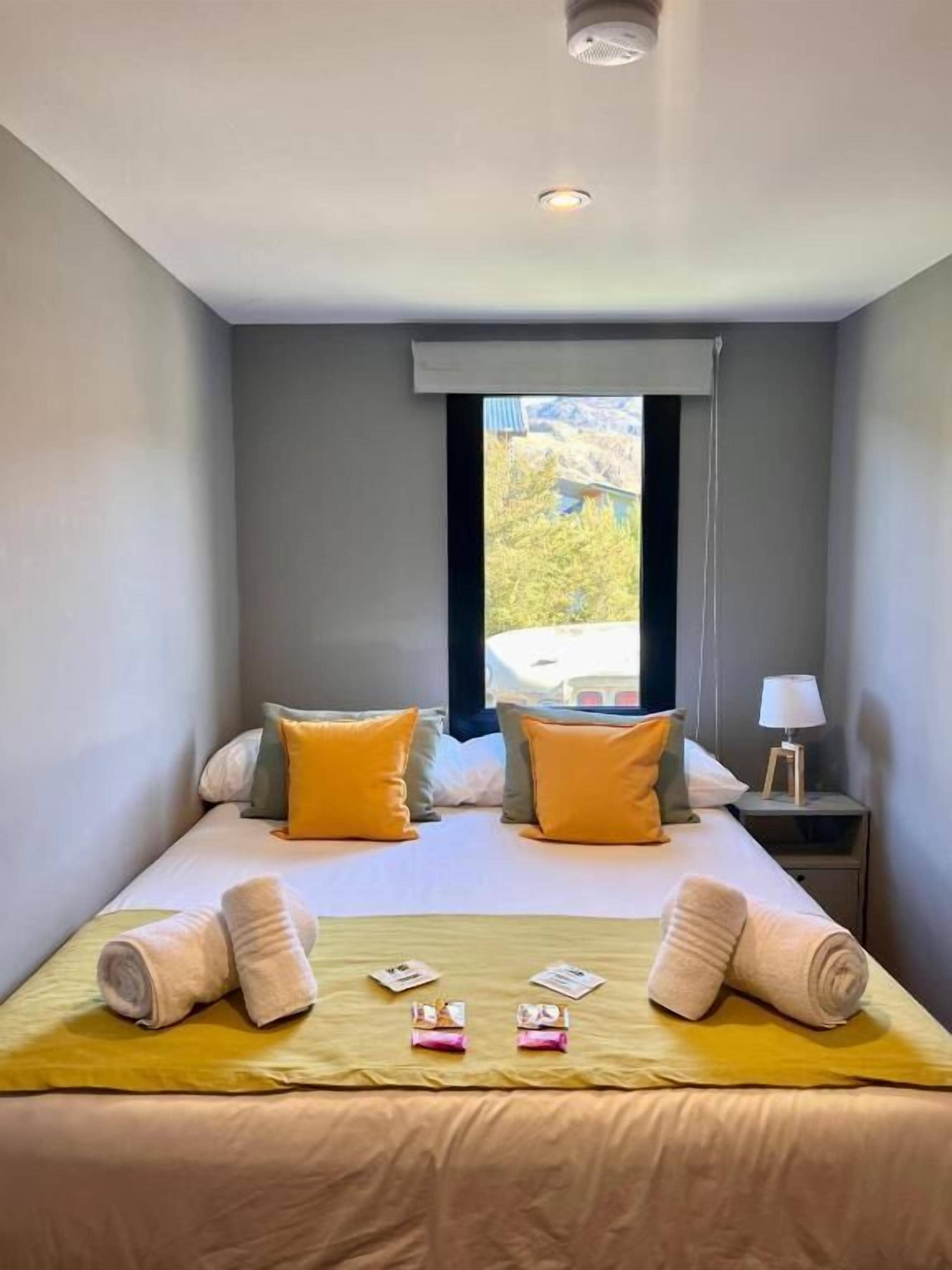 Habitación de hotel con cama, almohadas, mesita de noche, lámpara y ventana con vistas.