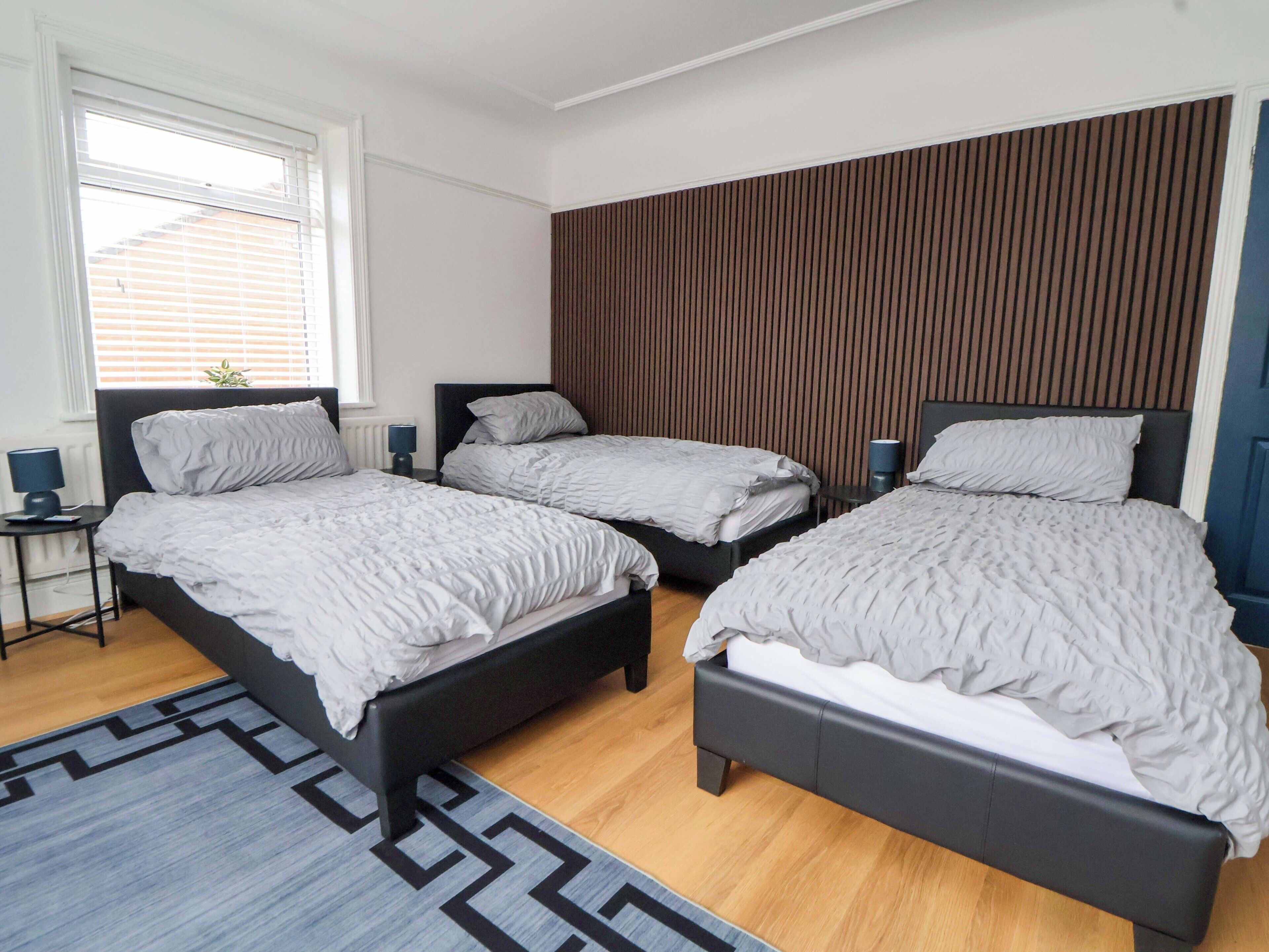 Un dormitorio con tres camas, una ventana con persianas, una mesita de noche con lámpara y una alfombra estampada.