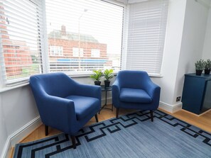 Une pièce avec deux fauteuils bleus, un tapis à motifs, une petite table avec une plante, et une vue sur un bâtiment en briques à travers de grandes fenêtres.