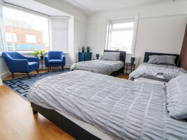 A bedroom with two beds, a blue armchair, a small table with a plant, and a view of a building through the window.