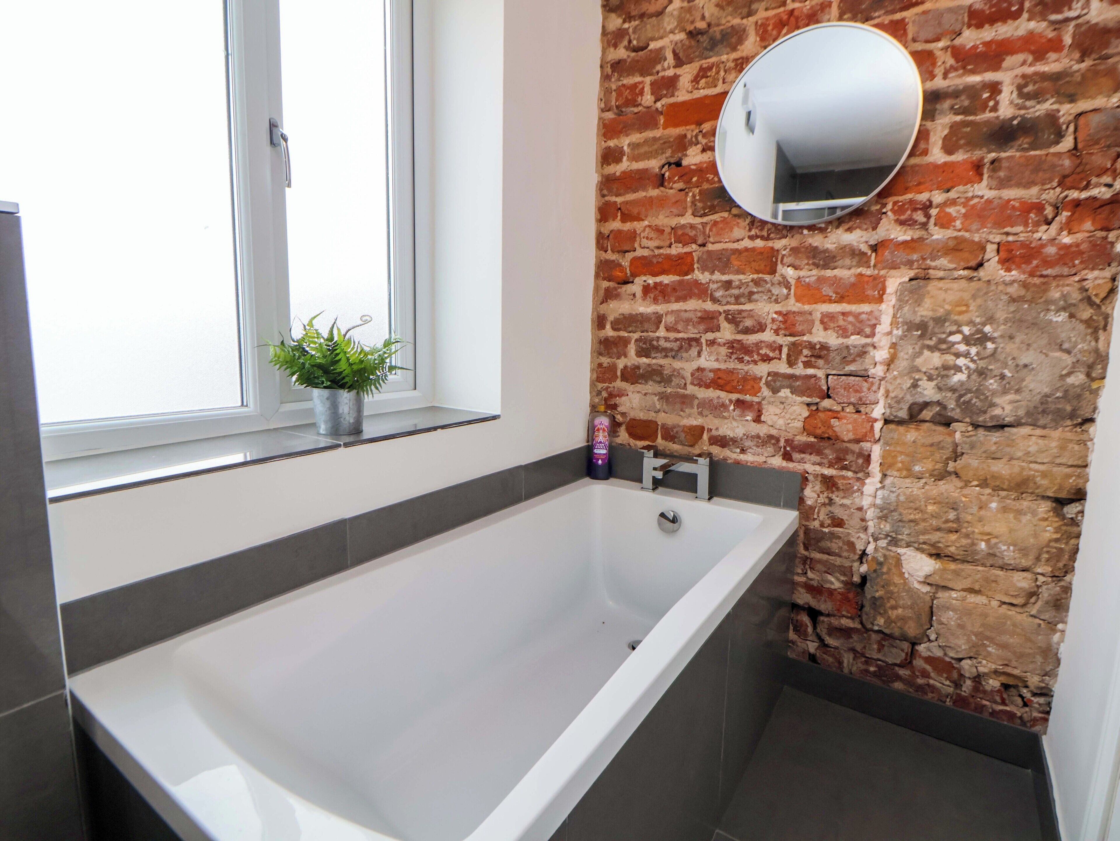 A bathroom with a large bathtub, a potted plant, a window, and a brick wall.