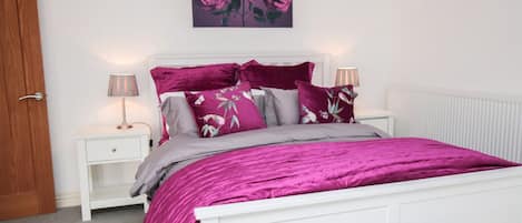 A bedroom with a white bed, purple bedding, and floral pillows.