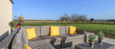 An outdoor seating area with a sectional sofa and a small table, surrounded by a wooden fence and a grassy field.