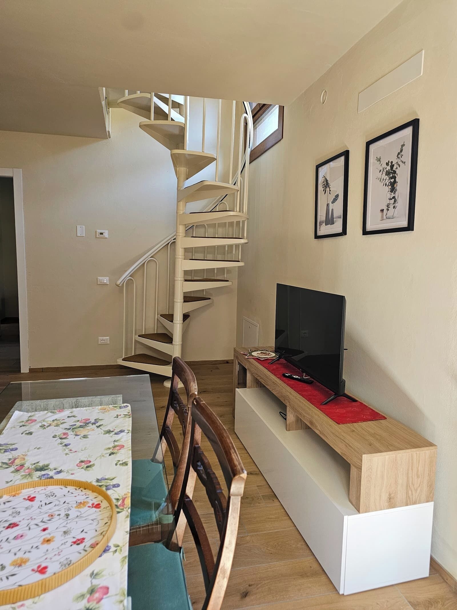 A modern living room with a wooden dining table, a TV on a wooden stand, a spiral staircase, and two framed pictures on the wall.