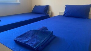 Zwei Einzelbetten mit blauer Bettwäsche, ein Fenster und ein an der Wand befestigter Nachttisch.