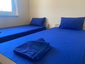 Zwei Einzelbetten mit blauer Bettwäsche, ein Fenster und ein an der Wand befestigter Nachttisch.