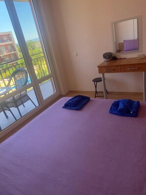 A room with a bed, a desk, and a chair. There is a balcony with a view of buildings and greenery. - Pool View 2 Bed at Holiday Fort Club (Nessebar)