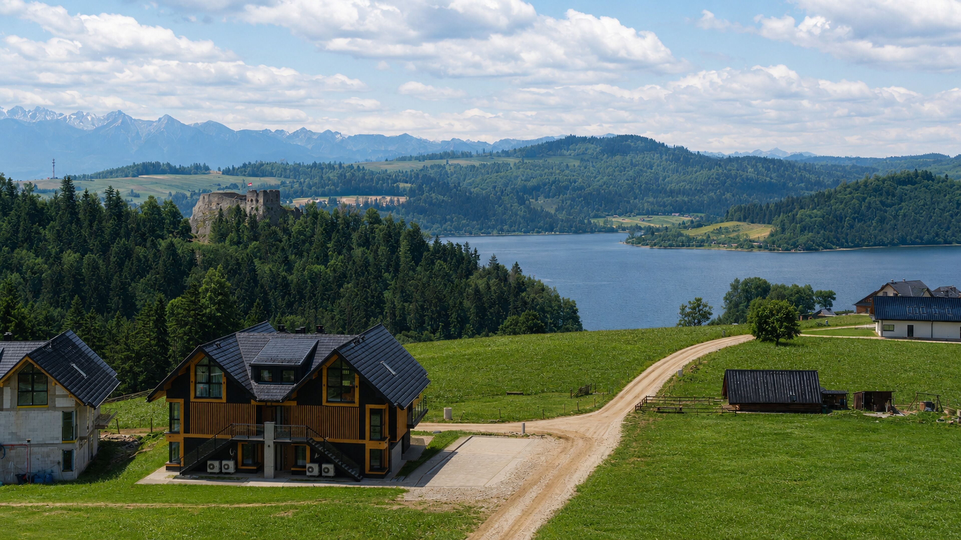 A wooden house with a paved driveway, surrounded by lush green fields and a lake.