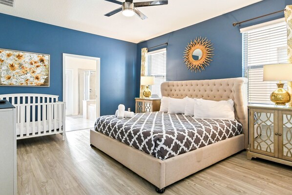 A bedroom with a tufted headboard bed, a crib, a sunburst mirror, and a wooden cabinet. - Spacious 9-bedroom house in serene Kissimmee with AC (Kissimmee)