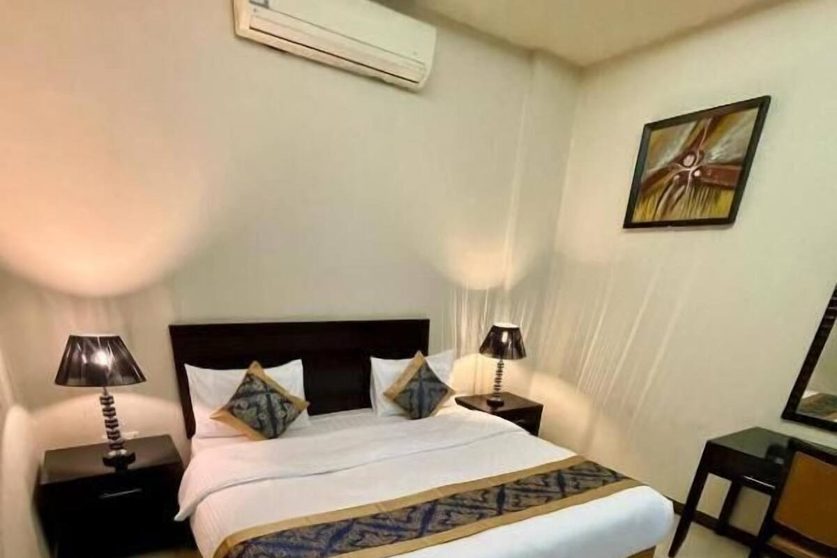 A neatly made bed with a patterned bedspread, two bedside tables with lamps, a desk, and a wall-mounted air conditioner.