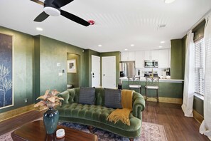 A living room with a green sofa, a coffee table, and a vase. There is a kitchen area with white cabinets and a microwave. - The Bavarian Inn 6 Chic 1 BDR (Covington)