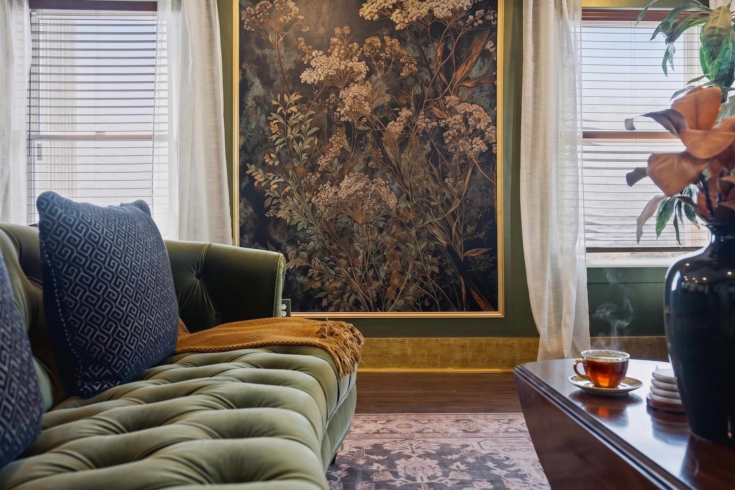 Een groene bank met een gekleurd kussen, een bijzettafeltje met kop en schotel, en een groot botanisch kunstwerk aan de muur.
