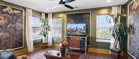 A living room with a ceiling fan, a cityscape painting, and large windows with curtains.