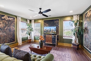 A living room with a ceiling fan, a cityscape painting, and large windows with curtains. - The Bavarian Inn 6 Chic 1 BDR (Covington)