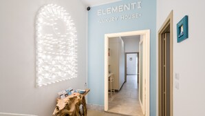 En hall med en väggskylt som visar texten ”ELEMENTI LUXURY HOUSE”, ett konstverk i trä och en dörr som leder till ett annat rum.