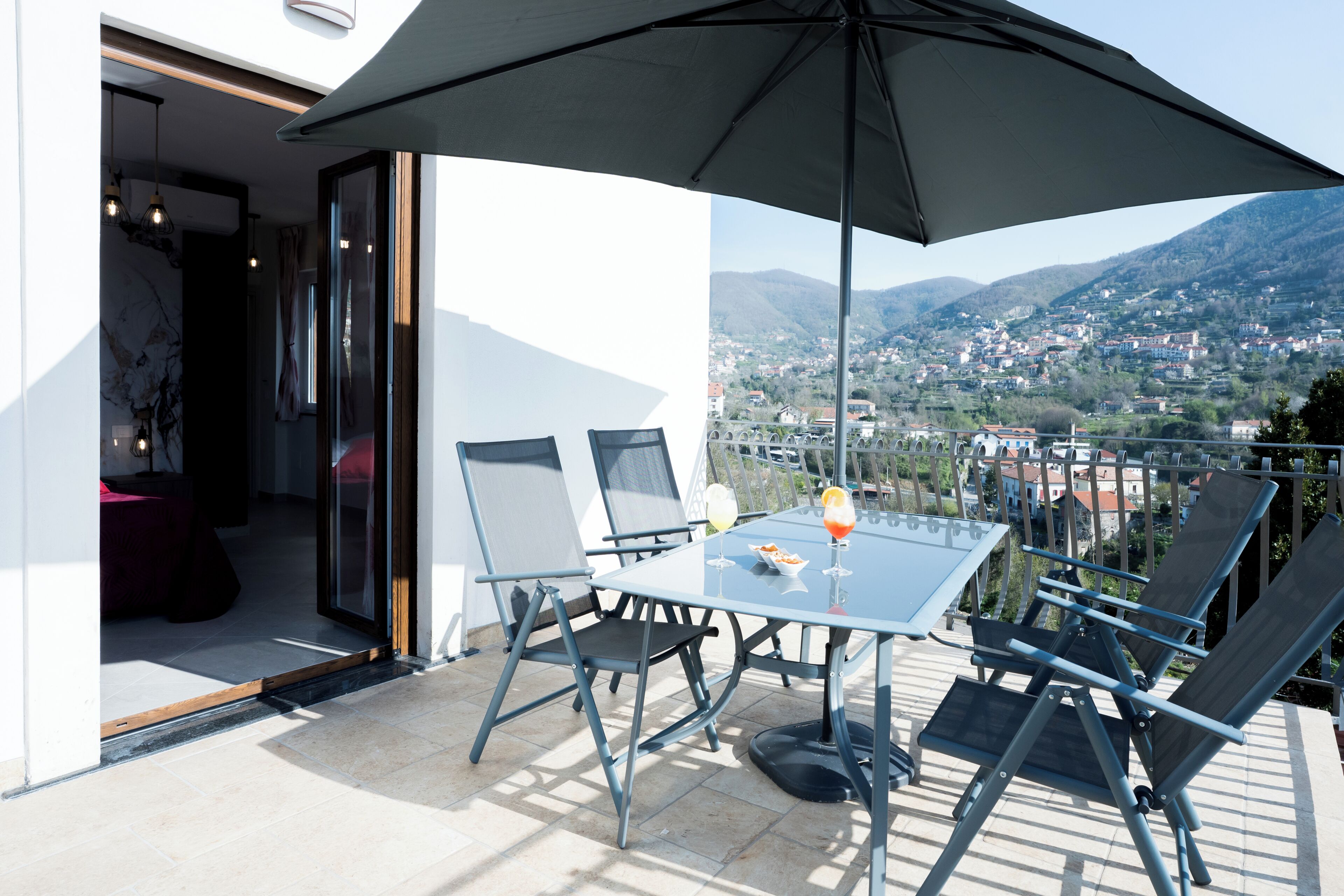 Una mesa de vidrio con sillas y una sombrilla en un balcón con vistas a un pueblo montañoso.