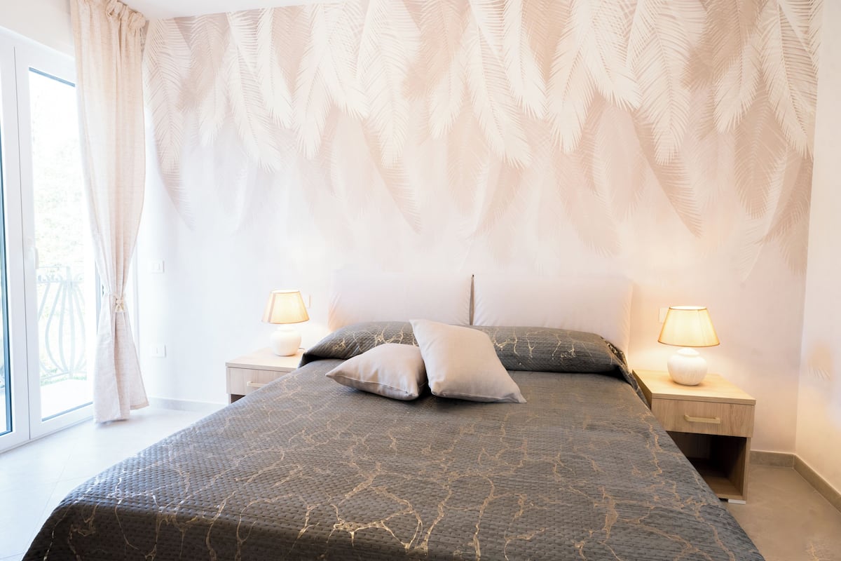 Une chambre à coucher avec un lit, deux tables de chevet avec des lampes, une fenêtre avec des rideaux et un mur orné d’un motif de feuilles.