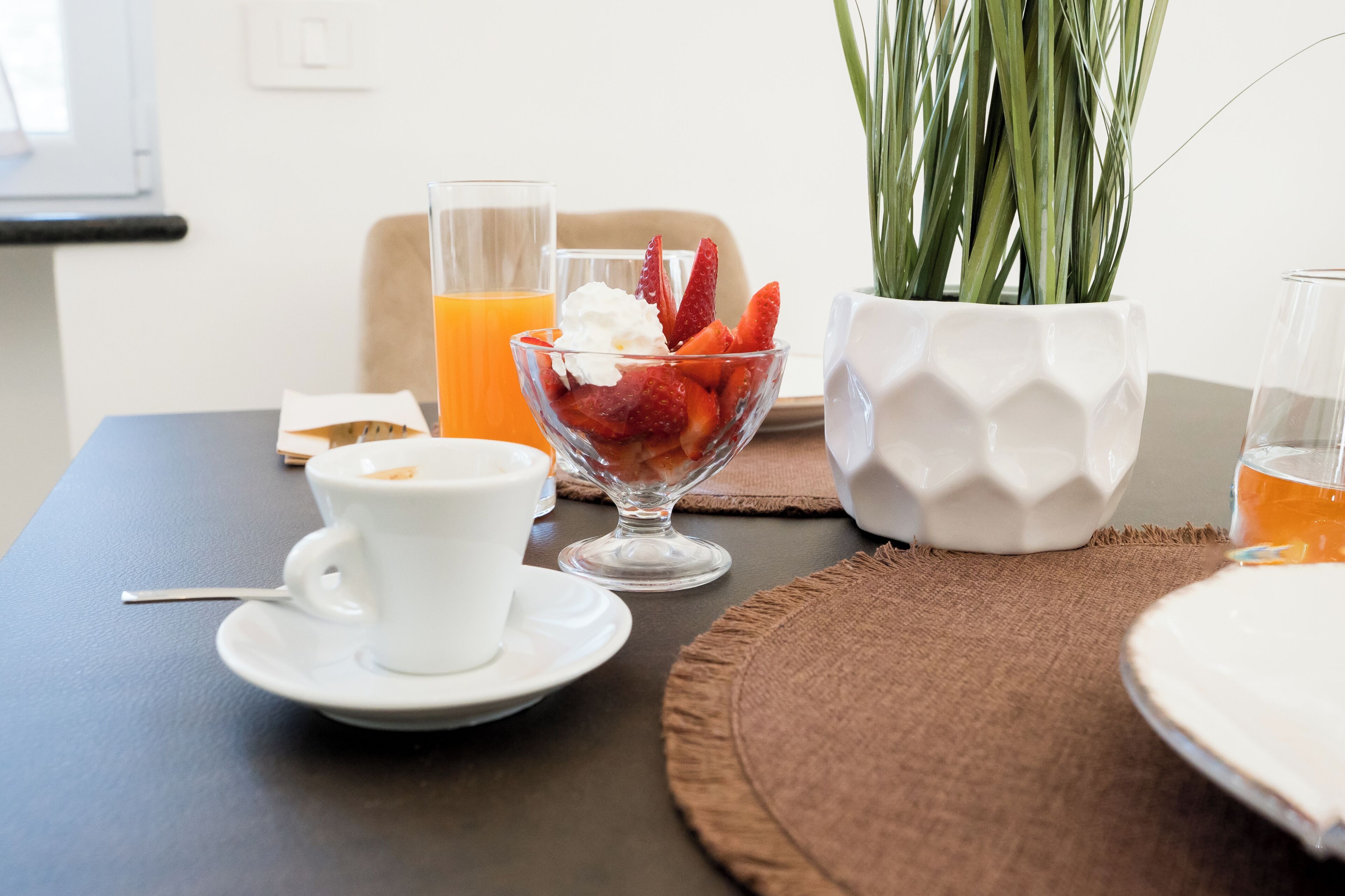 A breakfast setting with a glass bowl of strawberries and whipped cream, a glass of orange juice, a white cup, and a white pot with green plants.