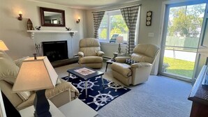 A living room with a fireplace, two armchairs, a sofa, a coffee table, and a large window with curtains. - 24 Riverbank Road. Footbridge Beach Cape! (Ogunquit)