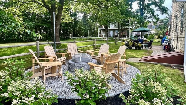 A patio area with Adirondack chairs around a stone fire pit, surrounded by gravel and greenery. - 24 Riverbank Road. Footbridge Beach Cape! (Ogunquit)