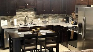 A modern kitchen with dark wood cabinets, a stainless steel refrigerator, a wall clock, and a decorative leaf wall art piece. - TinkerBell’s Smart Townhome  3BR & 3BA w/ a heated Pool 10 miles from Disney (Clermont)