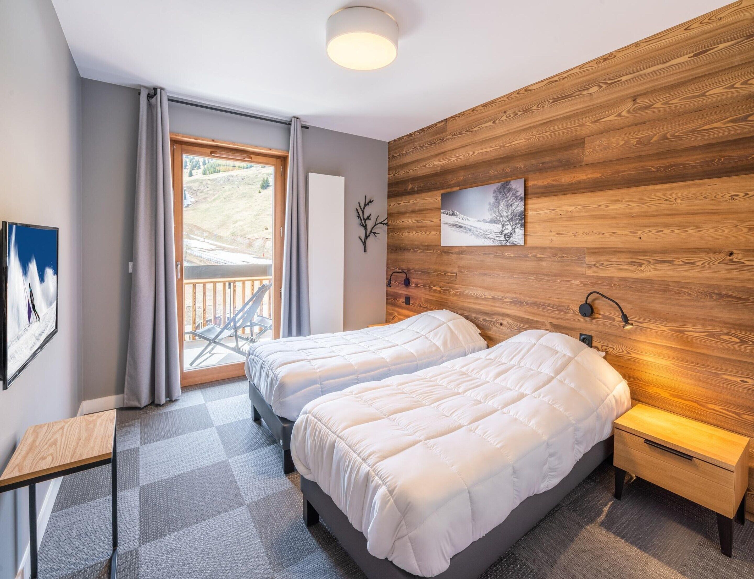 A bedroom with two beds, a wooden wall panel, a small table, a nightstand, and a balcony with a chair.