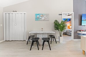 Zona pranzo con tavolo e sedie bianche, un quadro con conchiglie e stelle marine, una pianta e vista su una stanza con una TV.
