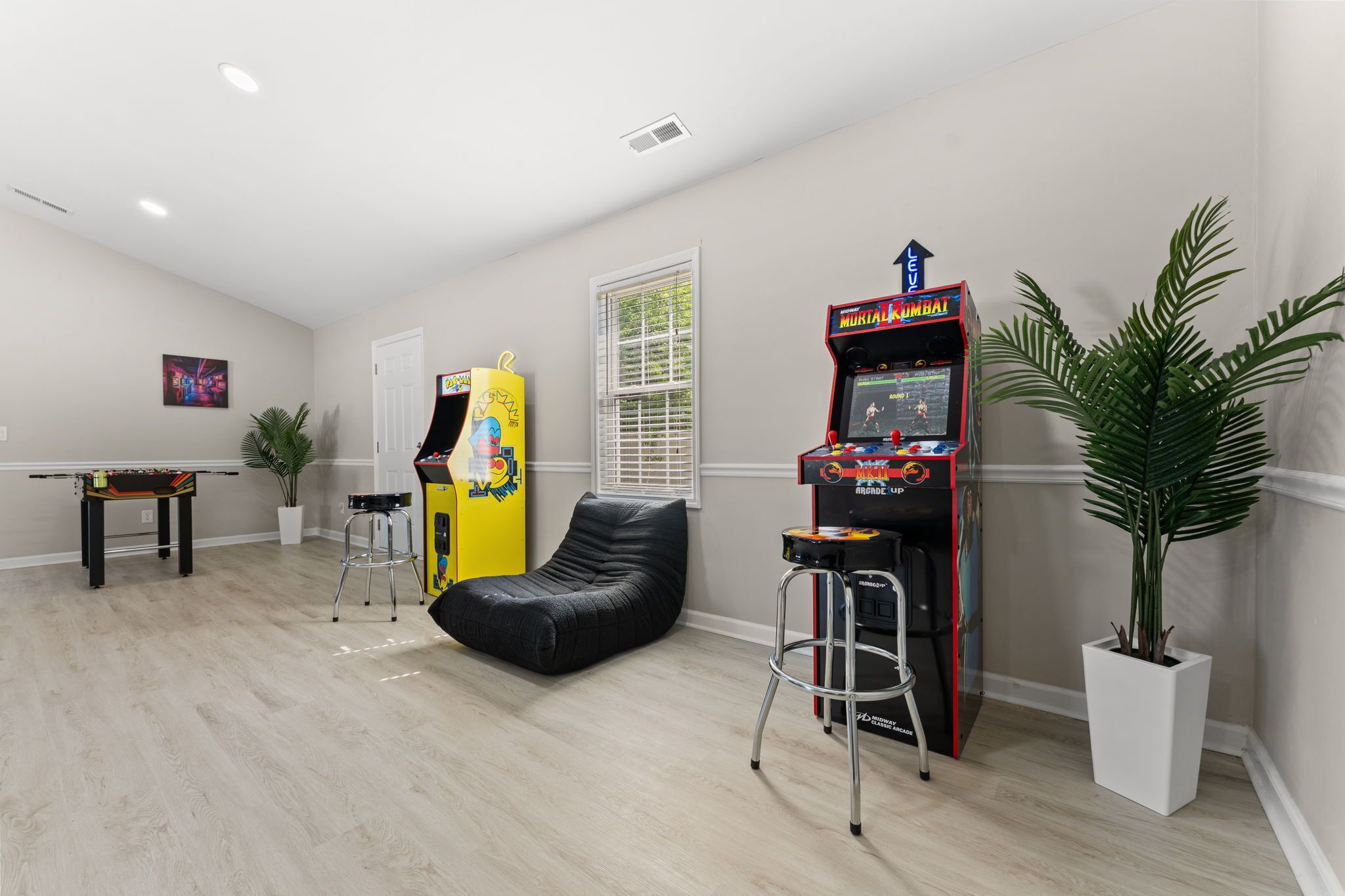 Um quarto com mesa de bilhar, uma máquina de jogos eletrônicos amarela, um pufe preto e uma máquina de jogos eletrônicos vermelha, além de uma planta em vaso.