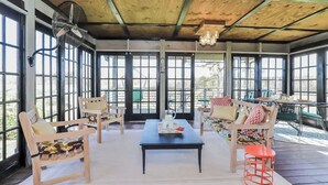 Un espacio de estar al aire libre cubierto, con mobiliario de madera, una mesa central y ventanas grandes.