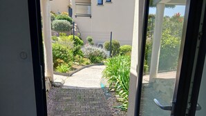 A view through a glass door into a courtyard with a paved walkway, a small garden, and a building in the background. - STUDIO sea and cape view 2pers. (Erquy)