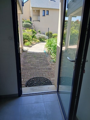 A view through a glass door into a courtyard with a paved walkway, a small garden, and a building in the background. - STUDIO sea and cape view 2pers. (Erquy)