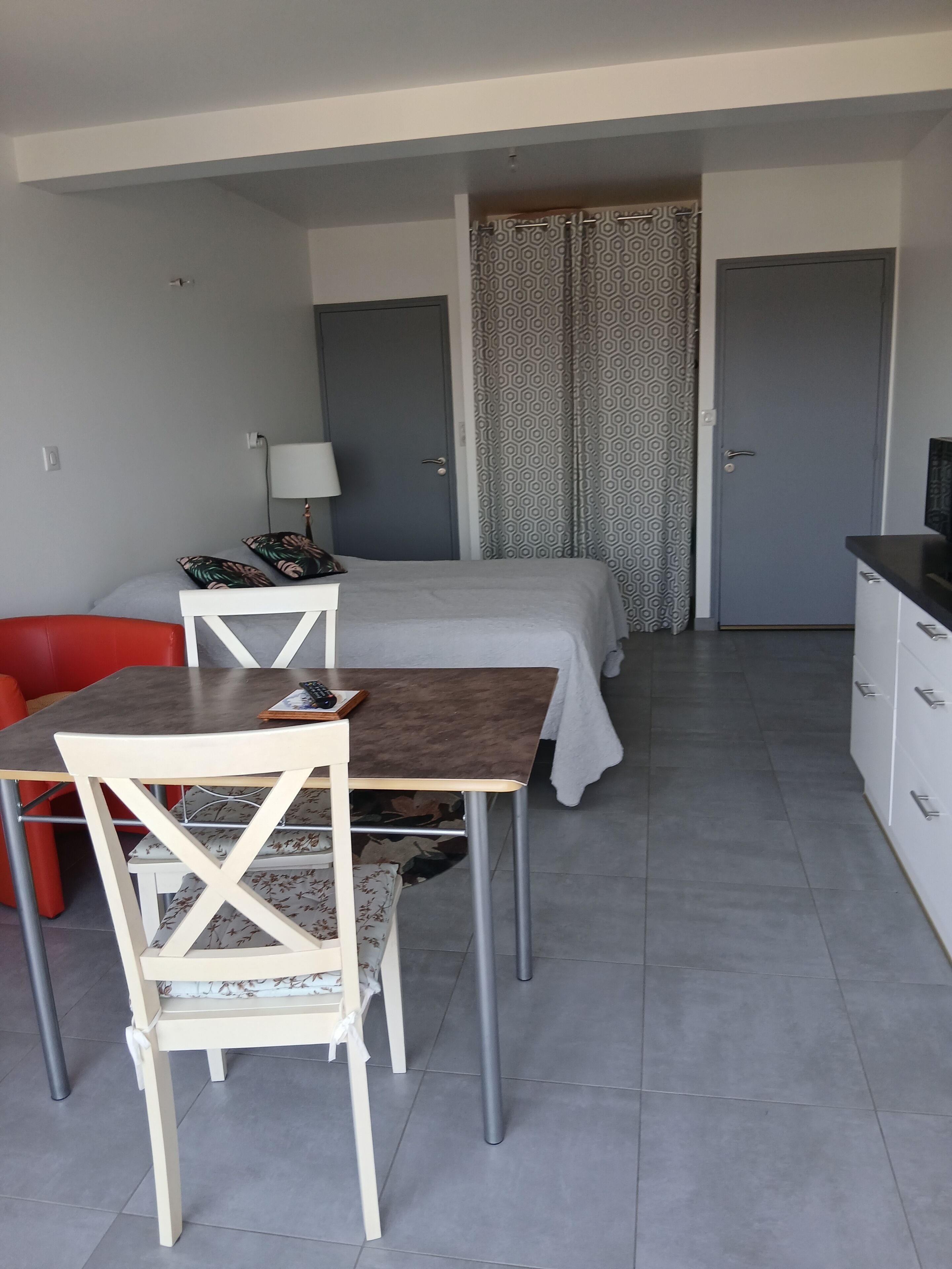 Quarto pequeno e compacto com uma cama, uma escrivaninha com abajur, uma cadeira e uma televisão.