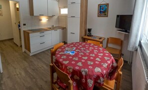 Una cocina compacta con armarios blancos, una mesa de comedor con mantel rojo y un televisor en la pared.