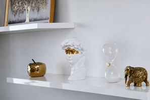 Uma estante branca com uma maçã dourada, um busto branco com detalhes dourados e uma figurinha de elefante dourado.