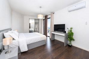 A modern hotel room with a bed, bedside lamp, TV, desk, and a potted plant.