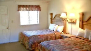 A room with two beds, a window with blinds, a door, and a nightstand with a lamp.