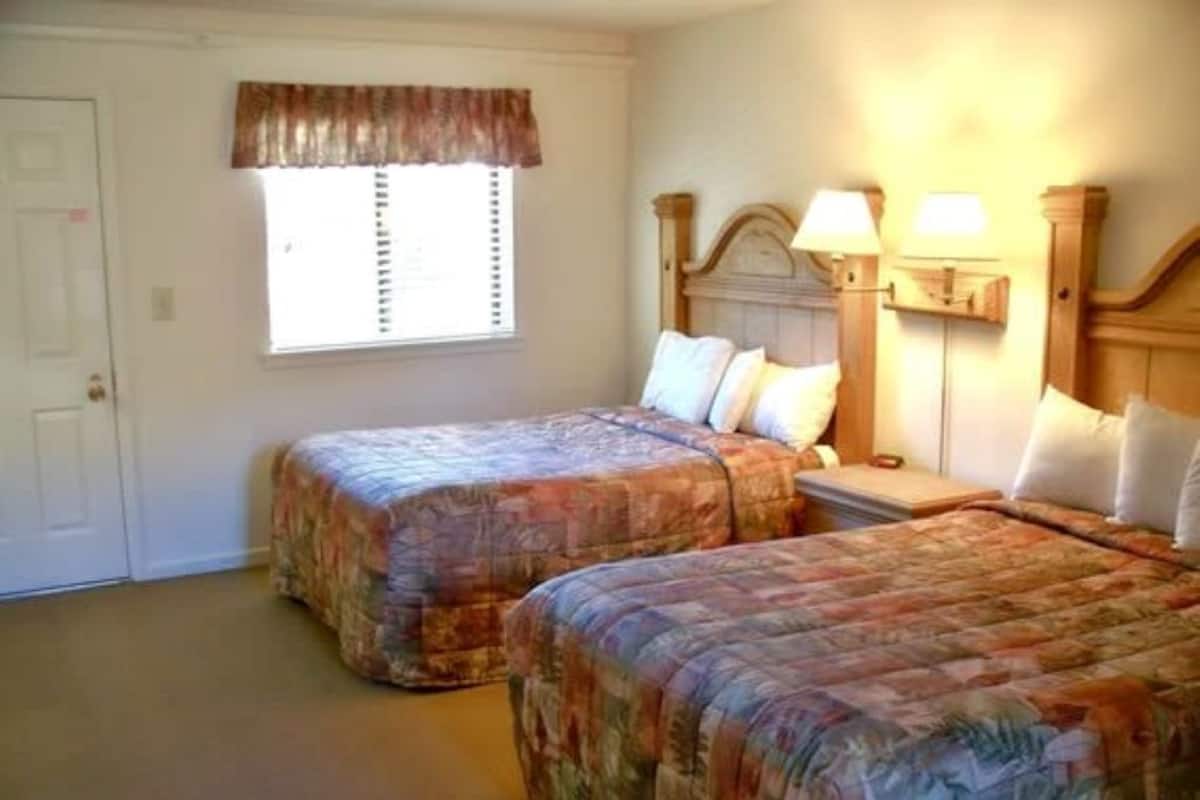 Una habitación con dos camas, una ventana con persianas, una puerta y una mesita de noche con lámpara.
