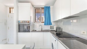 Une cuisine compacte avec des meubles blancs, un four encastré, un four à micro-ondes, un lave-linge et une fenêtre ornée d’un rideau bleu.