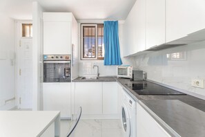 A compact kitchen with white cabinets, a built-in oven, a microwave, a washing machine, and a window with a blue curtain. - Feliz-Air-Los Cristianos Apartment (Arona)