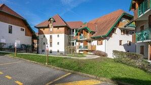 A row of traditional chalet-style buildings with wooden facades and pitched roofs, a clear blue sky, and a parking area with yellow lines. - Large 8-person Apartment in Giez (Giez)