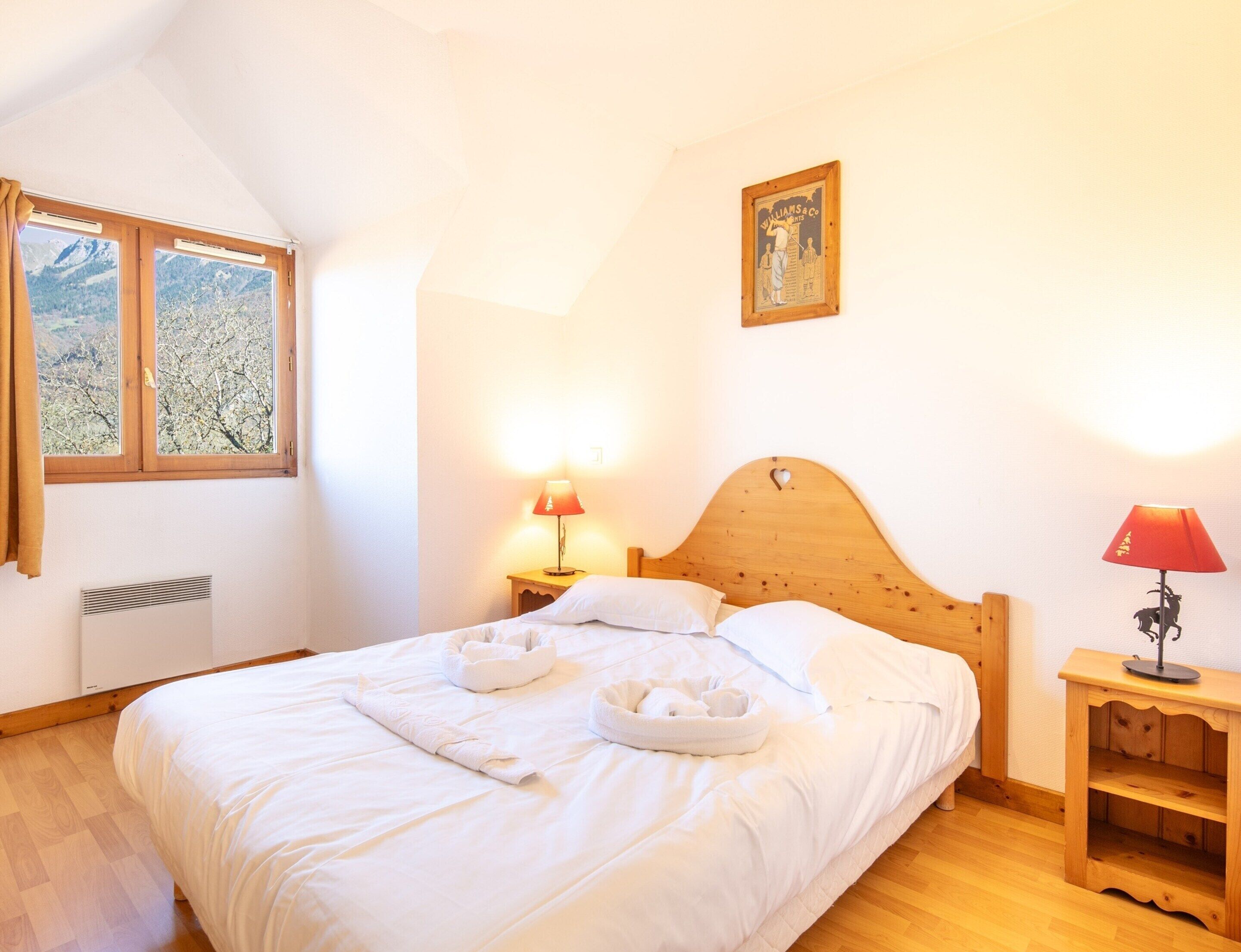 A bedroom with a wooden bed, two bedside lamps, a window with a view of trees, and a framed picture on the wall.