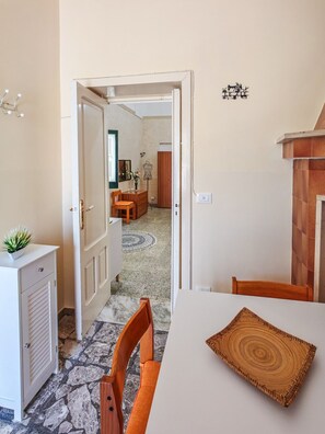 A dining area with a table, chairs, a decorative rug, and a view into another room with a wardrobe and a mannequin. - The House of Aunts - Maglie (Maglie)