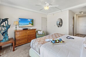 A bedroom with a bed, a ceiling fan, a television, a wooden dresser, a chair, and a painting of a beach. - Ocean Front steps from beach, dining, shopping 1051 (Lahaina)