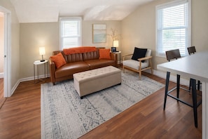 A living room with a brown leather sofa, a beige ottoman, a wooden chair, a dining table, and a patterned rug. - Lake Breeze Stylish Beach Boho 1BD w/ Private Deck (Michigan City)