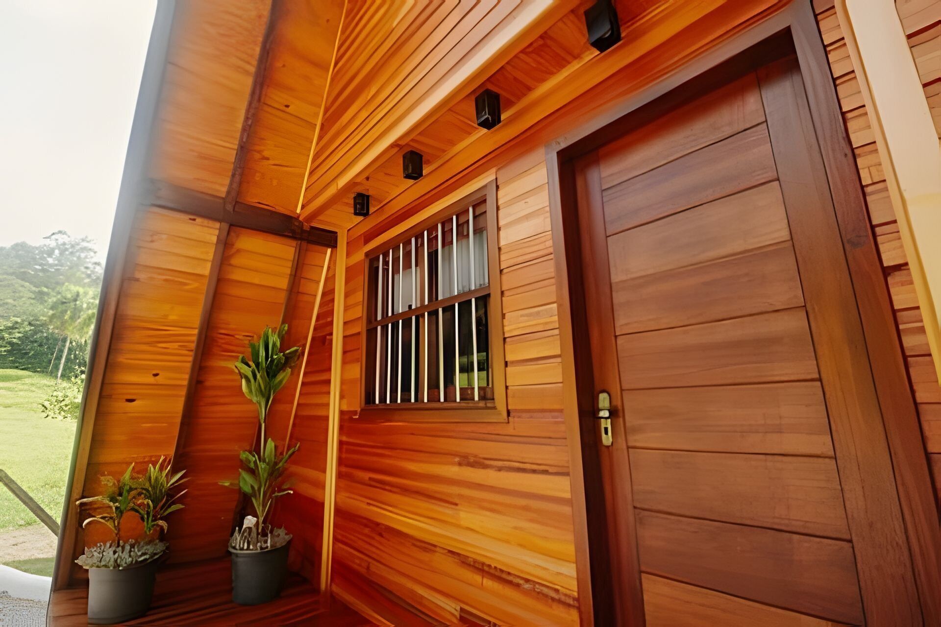 Een houten gebouw met een grote deur, een raam met tralies, en twee planten in potten op de veranda.