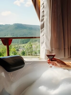 Un cuarto de spa con vista a un paisaje montañoso, una bañera y una mano sosteniendo una copa.